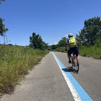 Cyclist riding along bike pathway marked with blue line in rural Japan with clear blue sky.