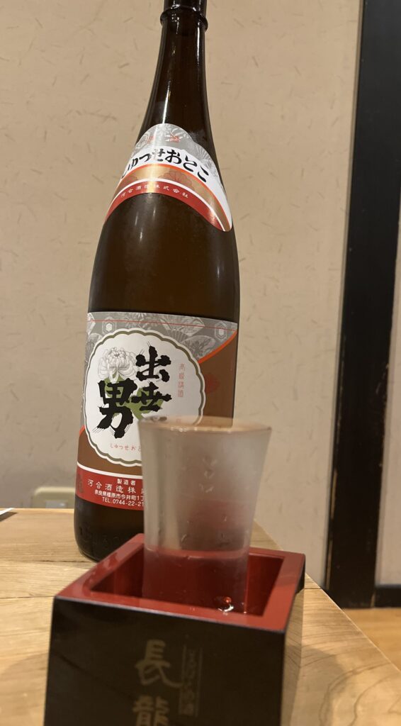 Bottle of large sake in background with a cup in a square dish serving the sake.