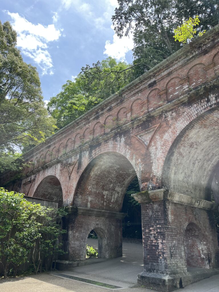 Large cement arches that have red discoloration due to their age with a bright blue sky and green trees in the background. Suirokau Aqueduct in Kyoto.