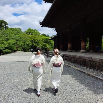 Two woman walk away in white kimonos with pink and red obis (belts) on a gravel path next to a temple.