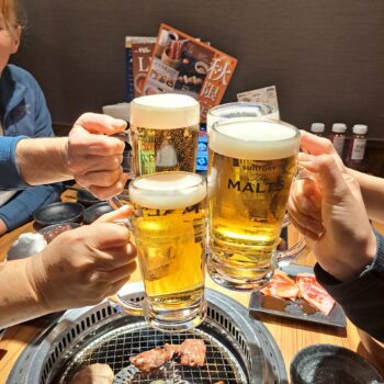 Four people toasting with large mugs of beer over a table with a yakiniku grill, featuring various meats and vegetables cooking, in a cozy restaurant setting with Japanese menus in the background.