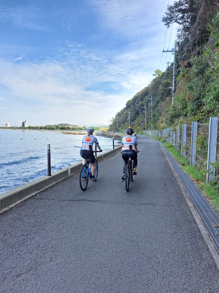 Two cyclist cycling away from the camera along a pathway along the ocean with green trees lining the right side of the pathway.