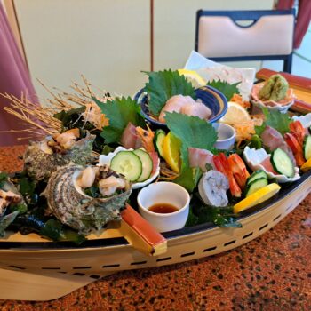 A small wooden boat serving fresh seafood including orange shrimp, seashells, tuna, lemon wedges, cucumbers.