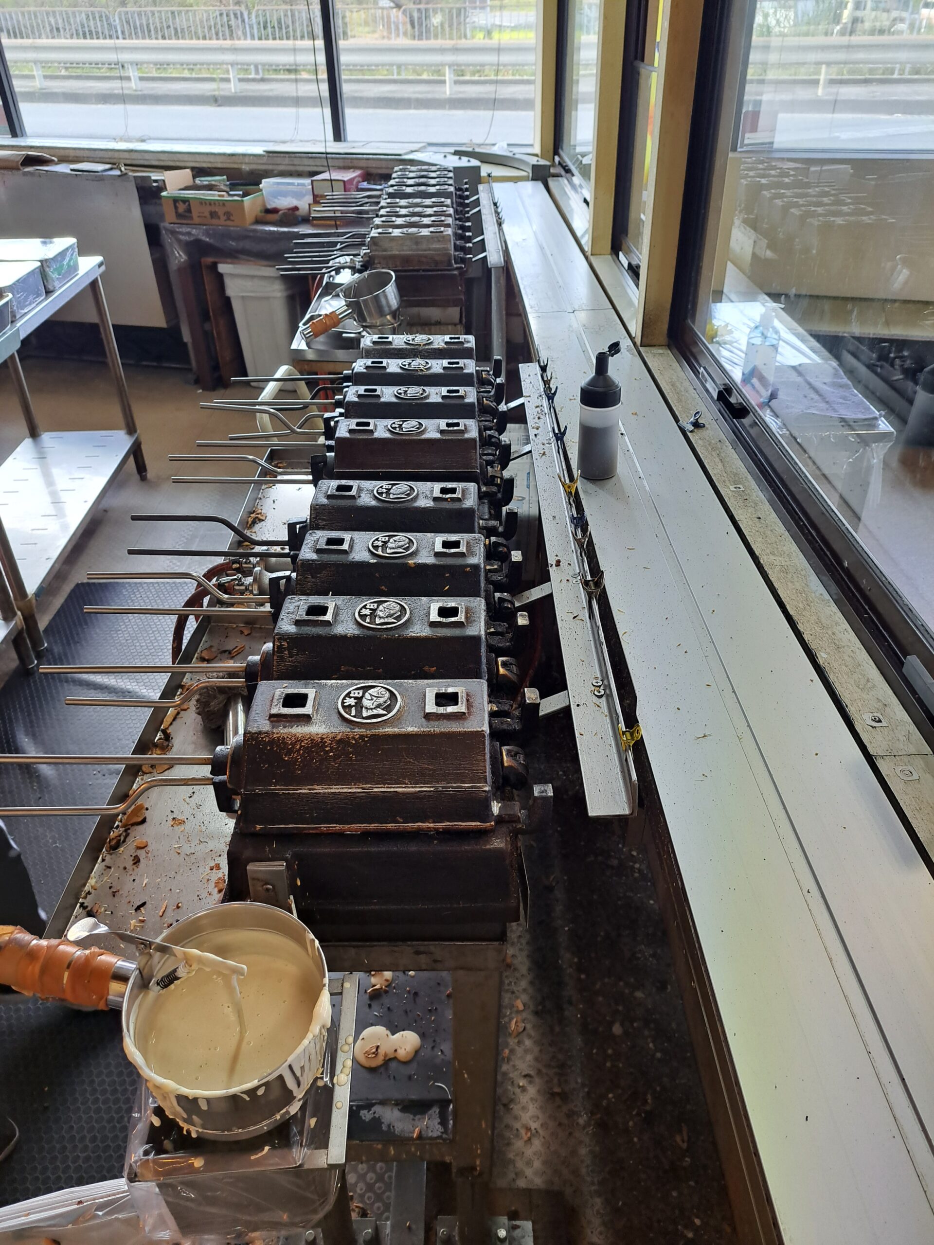 Making Taiyaki. A small pot of white sauce and a row of medal machines lined up ready to make the fish shaped sweet Japanese pastry filled with sweets.