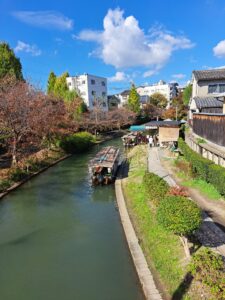 Water canal in the middle of the picture with two covered tourist boats , trees, green grass line the each side of the picture. A walkway is on the right side. A blue sky with a large white cloud is at the top of the picture.
