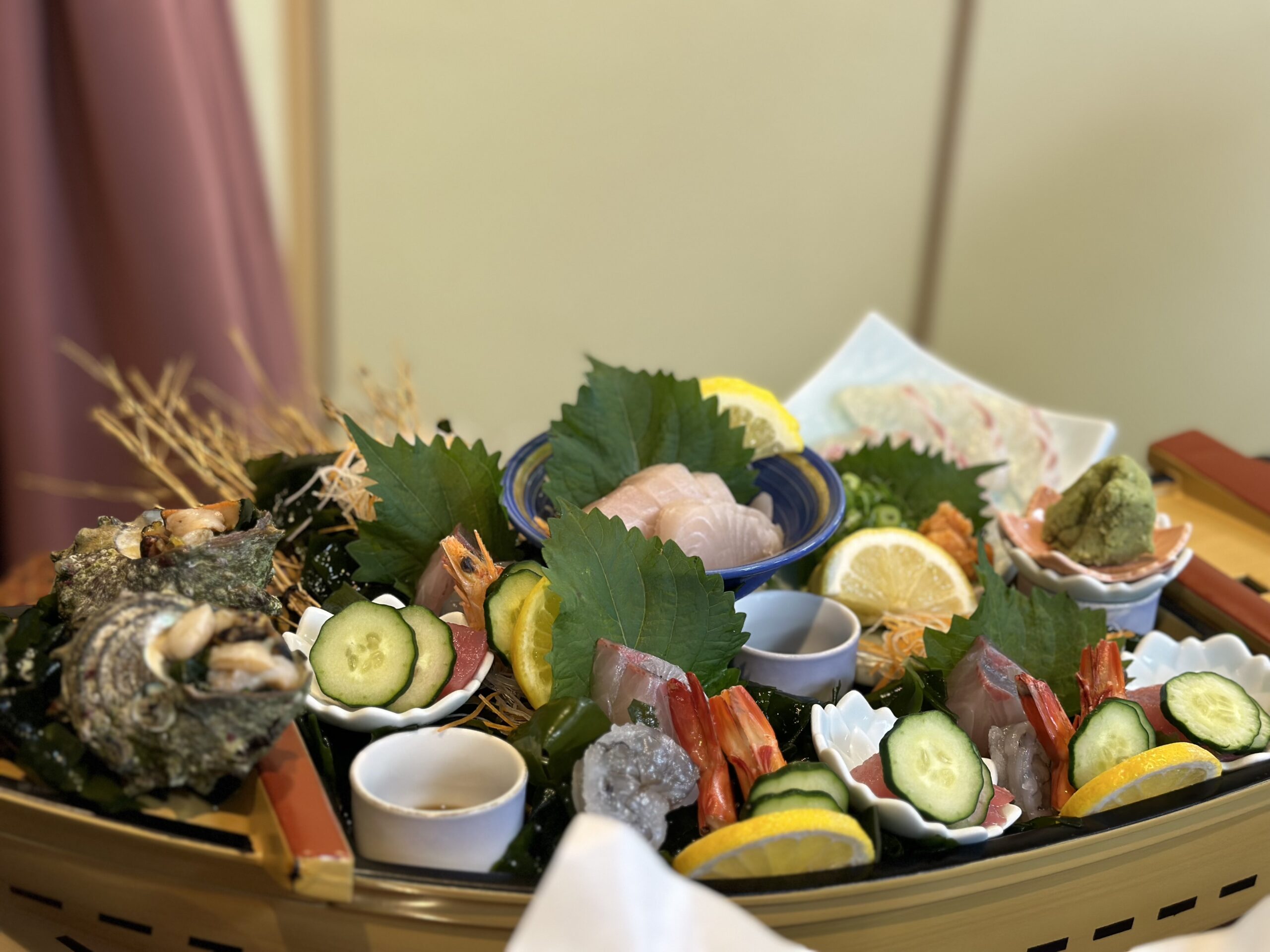 A decorative wooden boat filled with sashimi seafood including orange shrimp, dark seashells filled with the light fish meat, light colored tuna, with yellow lemon wedges and cucumbers between the dishes.