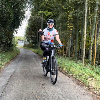 Cyclist waving as they bike on a paved path through a small bamboo grove.