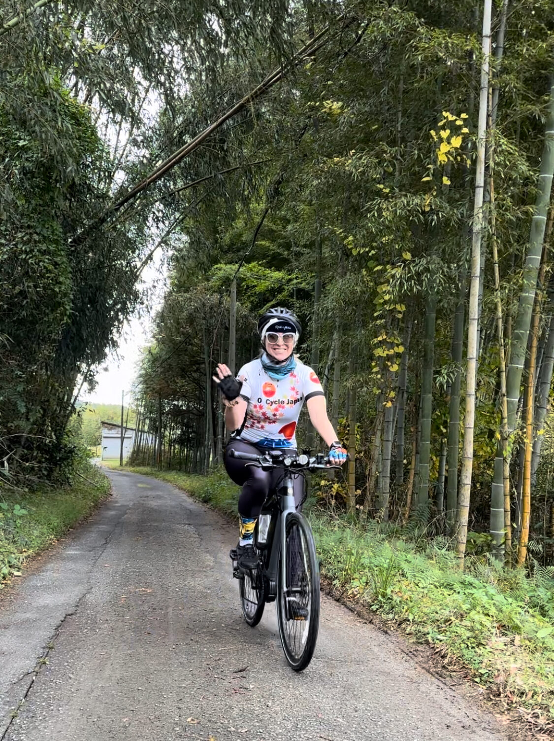 Cyclist waving as they bike on a paved path through a small bamboo grove.