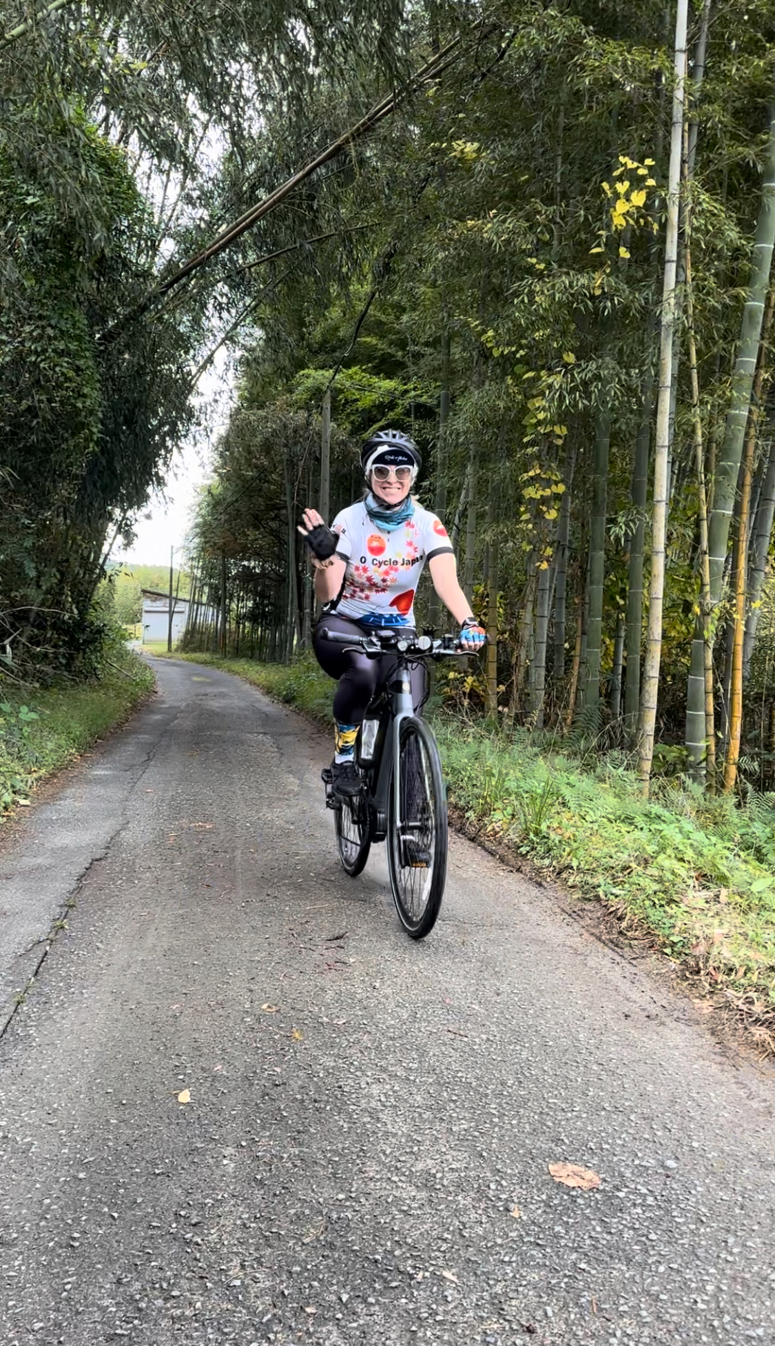 Cyclist waving as they bike on a paved path through a small bamboo grove.
