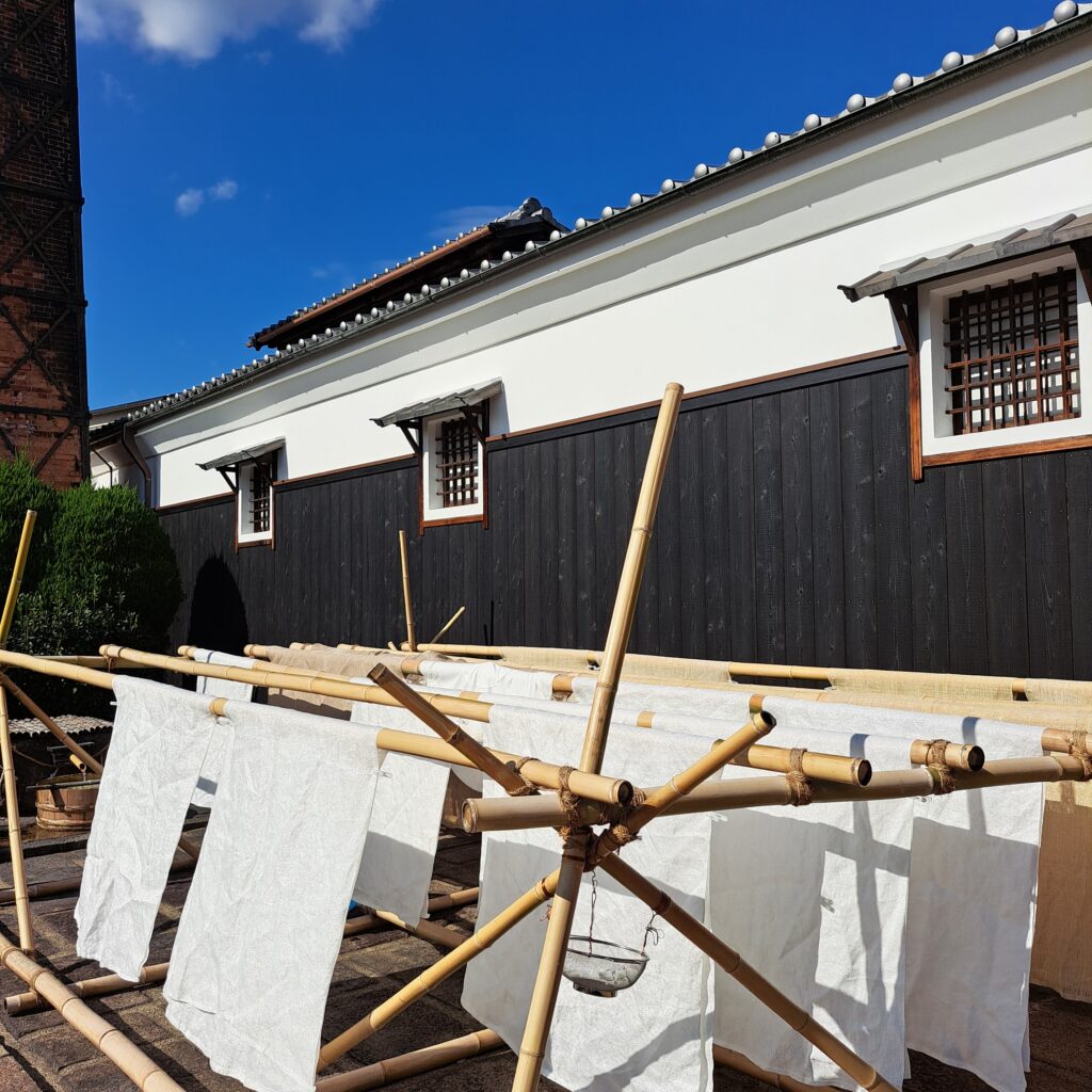 White clothes folded on bamboo stand in rows drying in a light breeze with brown and white Japanese building in background at the Gekkeikan Sake Museum.