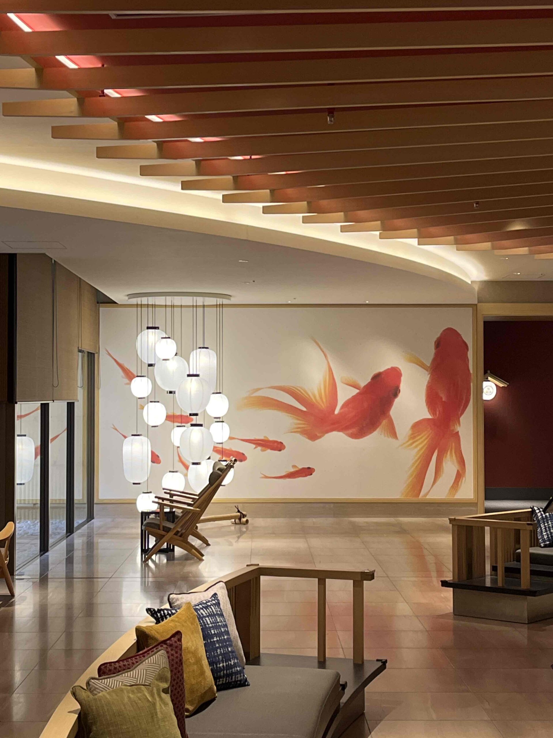 Japanese hotel in central Kyoto with large goldfish painted on the wall, hanging lamp display in the background and two lounge chairs. A bench with colored pillows faces the opposite direction.