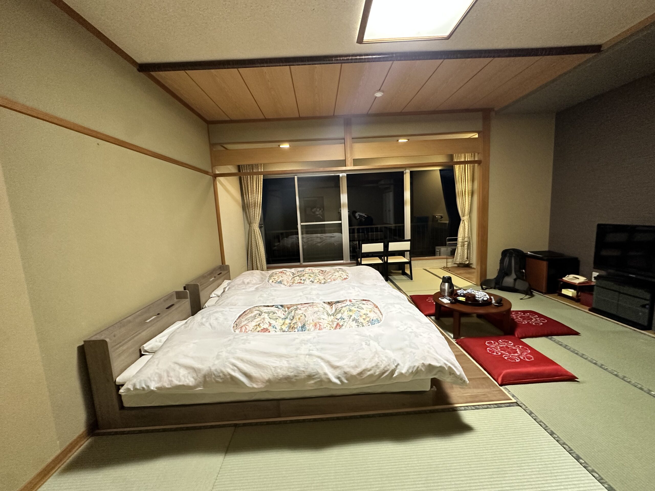 Japanese Roykan with western style bed frames, three red cushions around a low dark wooden table at the foot of the two beds. A large window is at the end of the room dark in the night.
