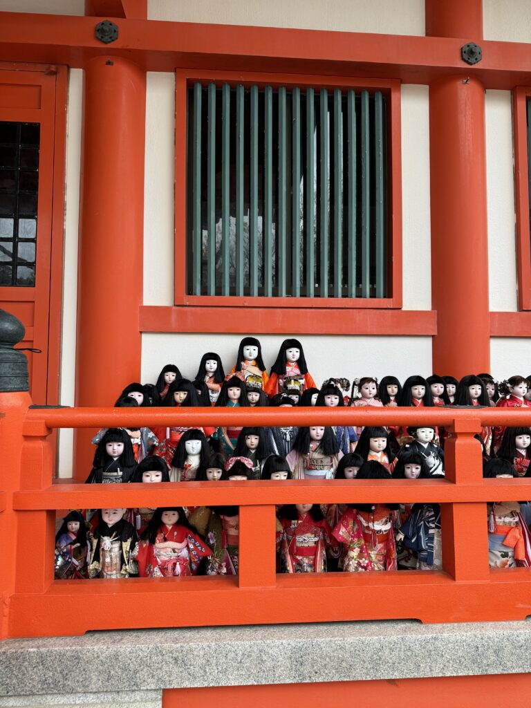 Layers of many Japanese dolls with white faces, black hair and traditional kimonos are lined up behind a red barrier in front of a shrine.