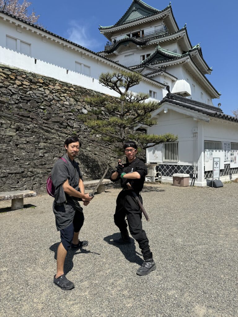 Two mend in dark clothing, one with a sword pose in front of a white castle.