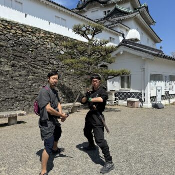 Two mend in dark clothing, one with a sword pose in front of a white castle.