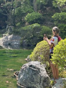 Person takes photo of a pond in a lush green Japanese garden.