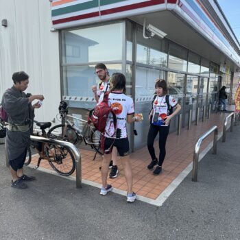 four people stand with two bikes at the corner of a convenient store with orange, green and red stripes on the building.