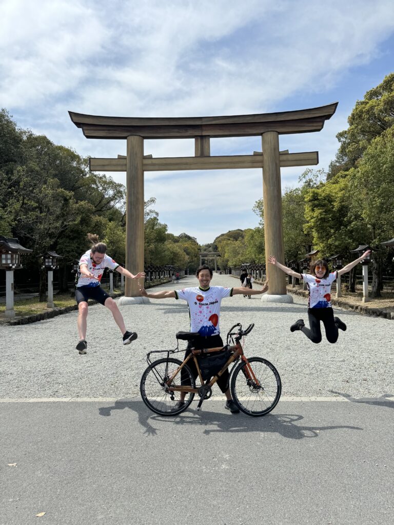 A man stands behind a bicycle with his hands outstretched as two people on either side are in the air. There is a large Tori gate in the background.