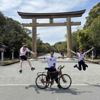 A man stands behind a bicycle with his hands outstretched as two people on either side are in the air. There is a large Tori gate in the background.