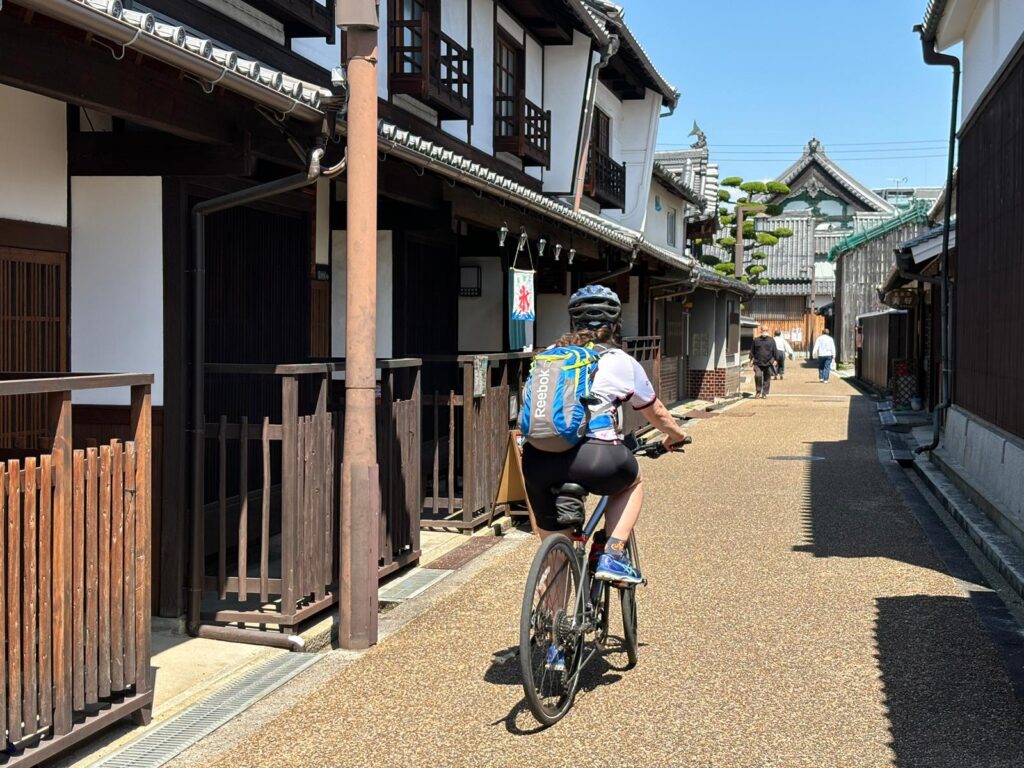 Cyclist riding through old Japanese neighborhood called Imaicho. White buildings with dark wood storefronts.