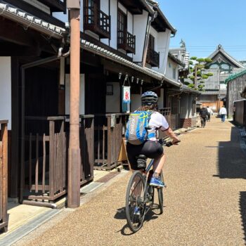 Cyclist riding through old Japanese neighborhood called Imaicho. White buildings with dark wood storefronts.