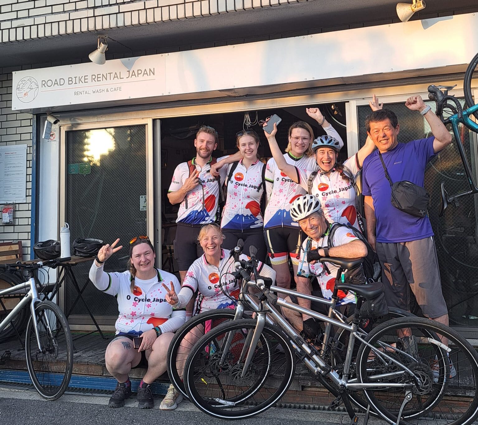 Group of seven cyclists in matching O Cycle Japan jersey's with man in Blue shirt in front of Road Bike Rental Japan Osaka office with bikes.