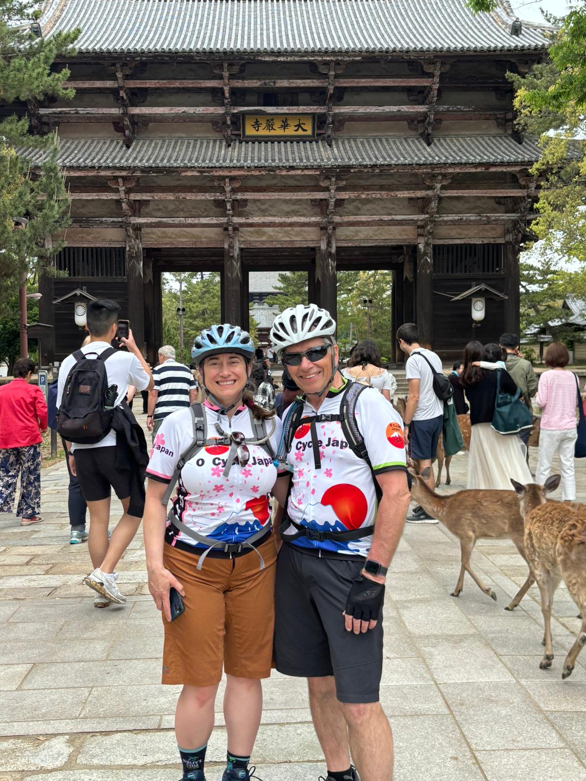 Two cyclists pose in front of temple entrance with deer walking in background.