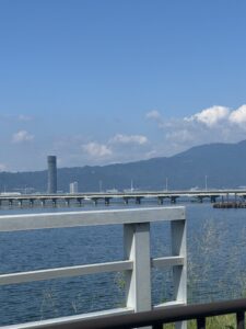 Blue water of Lake Biwa from the shore, with a bridge and tall building in the background.
