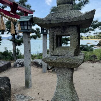 An Ishidoro or Japanese stone lantern is in the foreground on a beach with a lake and a red tori gate in the background.