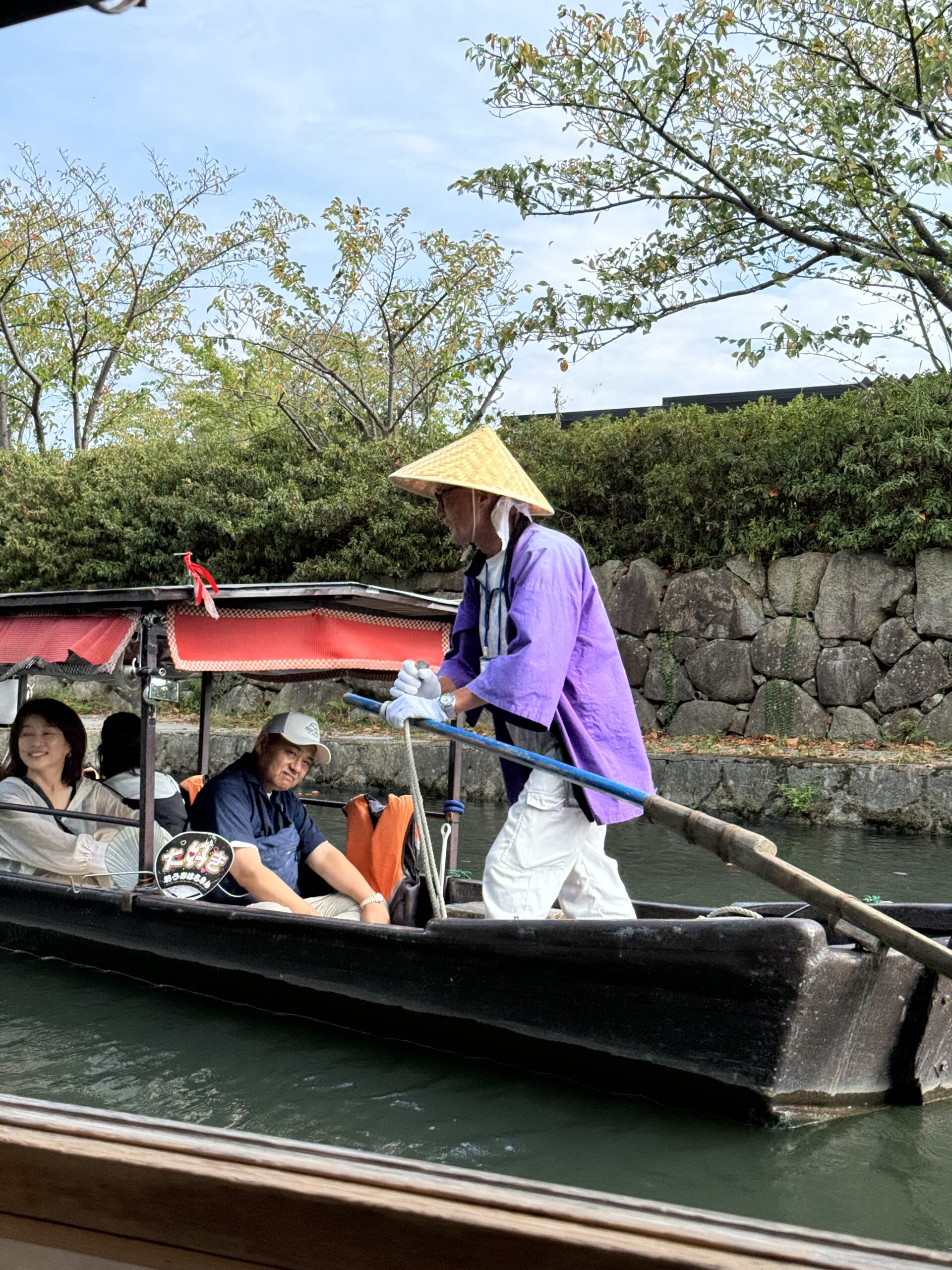 Man in purple goat and triangle straw hat is at the head of a boat full of tourists going done the canal at Omihachiman.