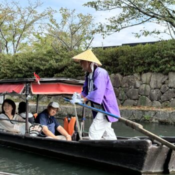 Man in purple goat and triangle straw hat is at the head of a boat full of tourists going done the canal at Omihachiman.