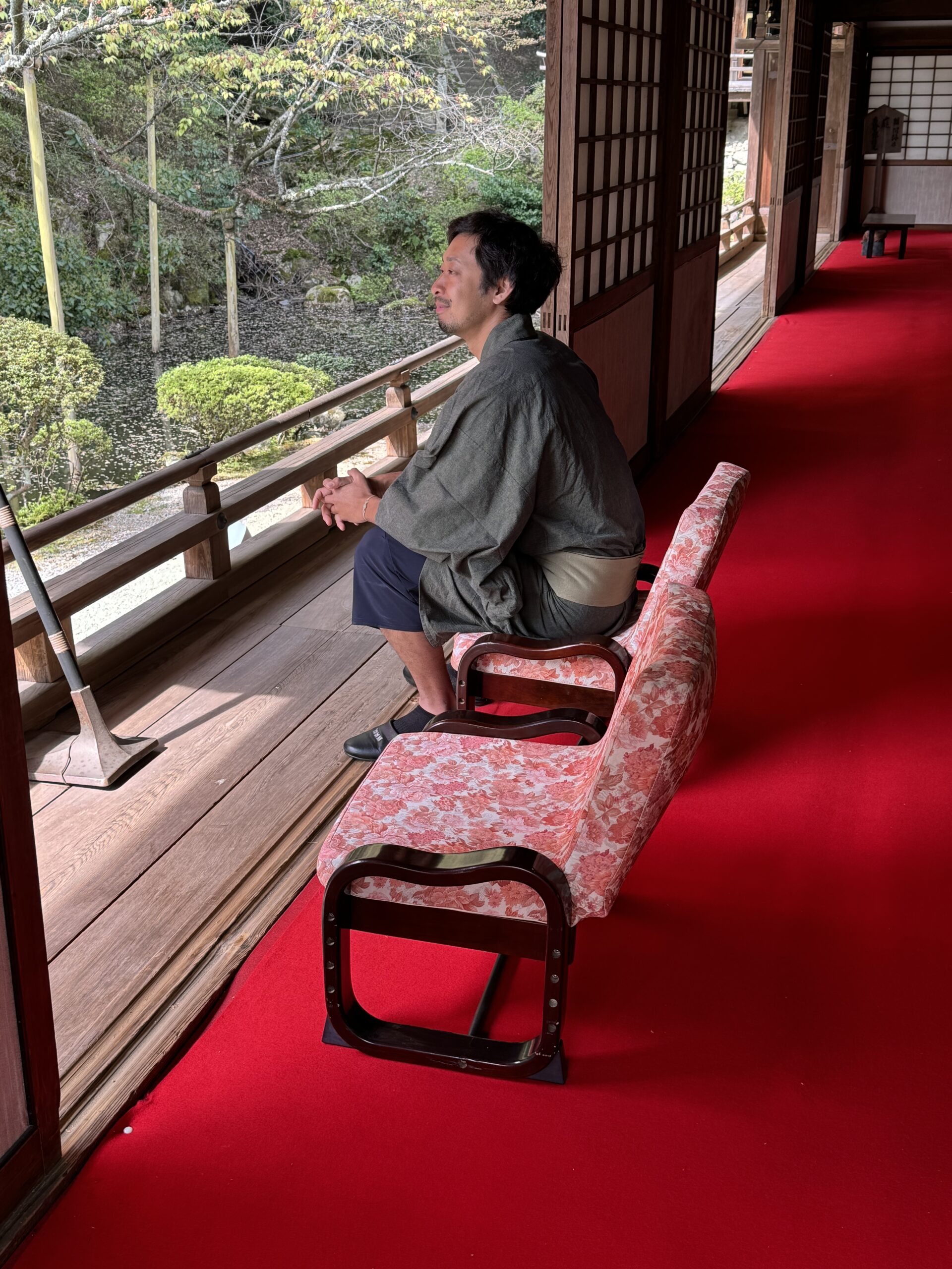 Man dressed in traditional Japanese clothes sits on a chair admiring a Japanese garden at a Monzeki.