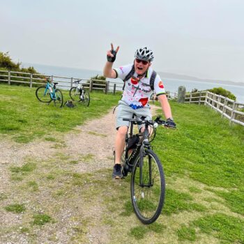 Cyclist gives piece sign as they ride towards the camera in gravel path. Two bikes are in the background. There isa fence and a blue-grey ocean and sky in the background.