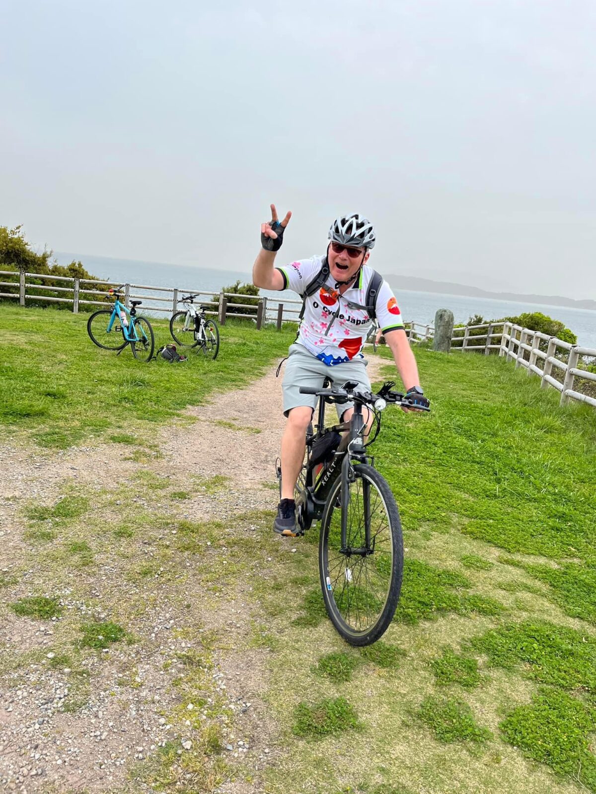 Cyclist gives piece sign as they ride towards the camera in gravel path. Two bikes are in the background. There isa fence and a blue-grey ocean and sky in the background.
