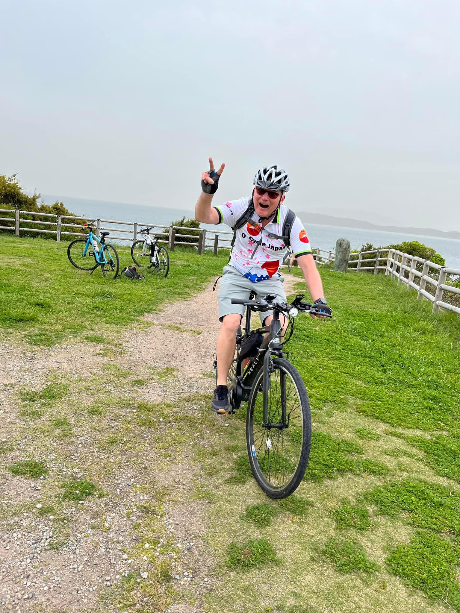 Cyclist gives piece sign as they ride towards the camera in gravel path. Two bikes are in the background. There isa fence and a blue-grey ocean and sky in the background.