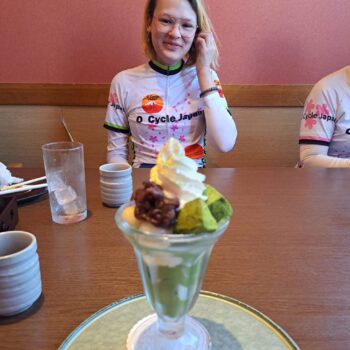 Cyclist poses with large fruit parfait dessert in front of her at a restaurant.