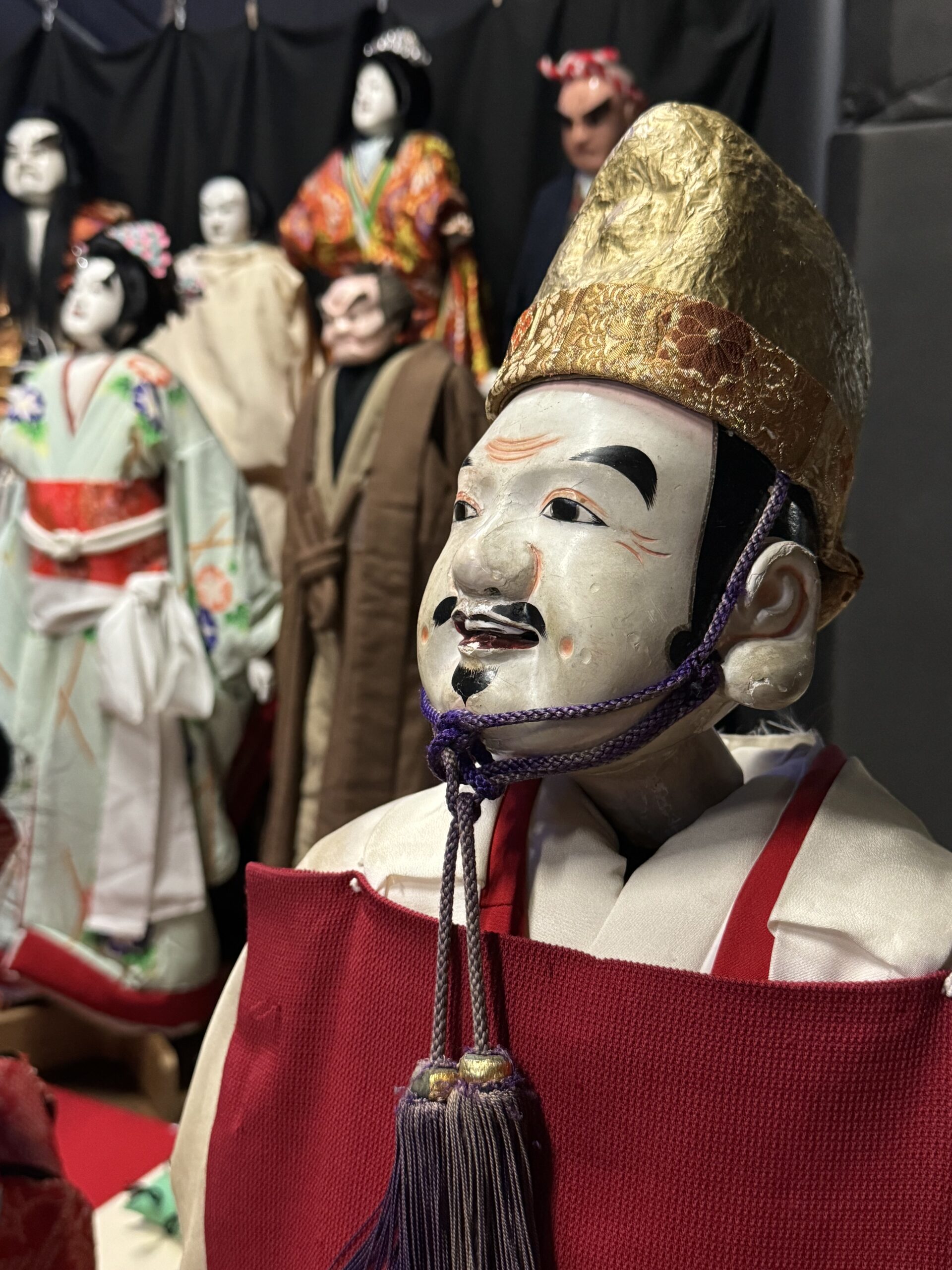 Puppet image of Ebisu, Japanese god of fisherman and luck. Gold had and red top with other wooden puppets in the background.