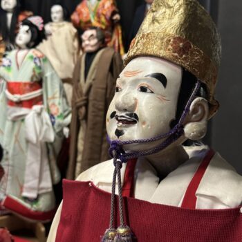 Puppet image of Ebisu, Japanese god of fisherman and luck. Gold had and red top with other wooden puppets in the background.