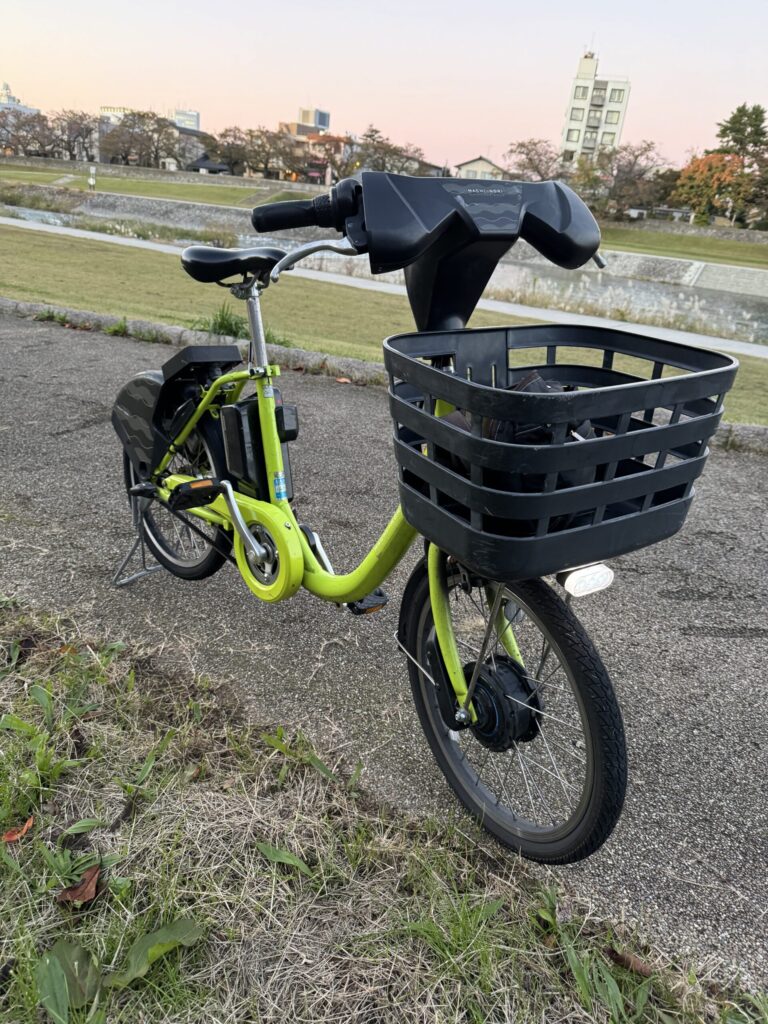 A lime green E-bike with large front basket on pathway in Kanazawa with light pink sky as dusk approaches.