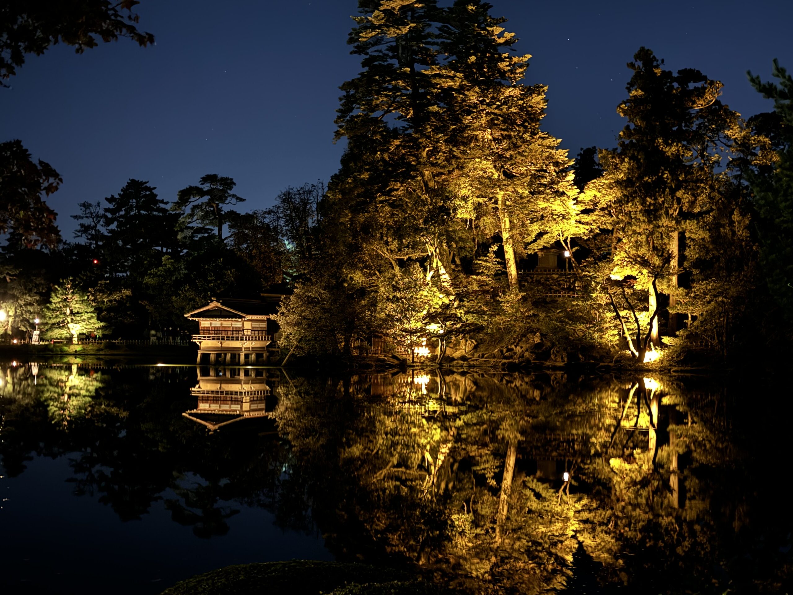 Kenrokuen Garden at night in Kanazawa. A small wooden Japanese structure sits at the waters edge near large cedar trees in a yellow glow reflected in the pond.