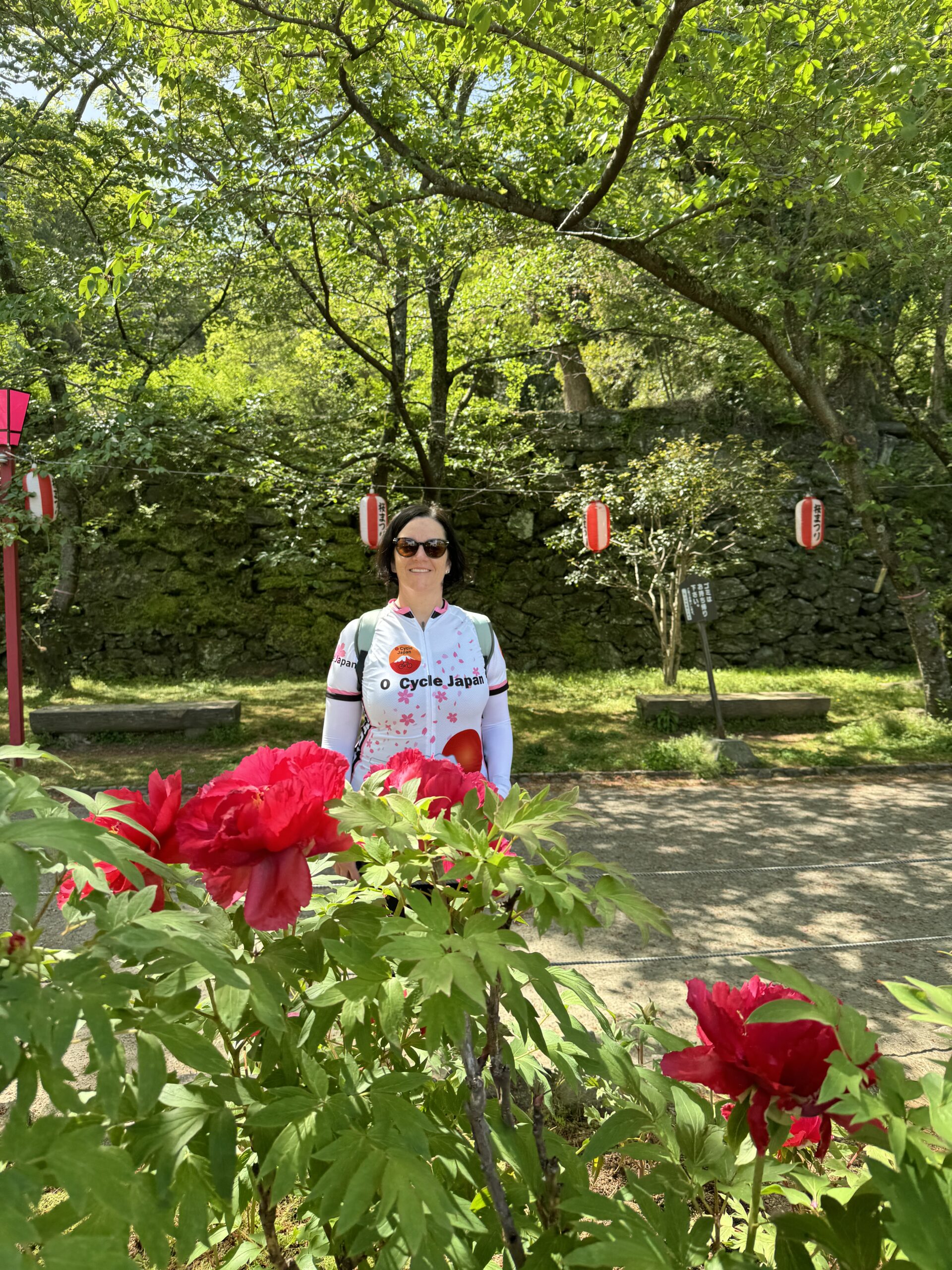 Discover Japanese Gardens. Woman stands behind red blossoms with red and white lanterns in the background.