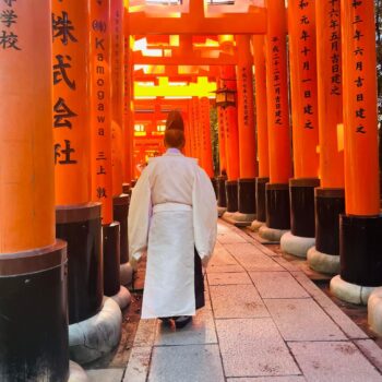 Buddhist priest at Fushimi Inari. Large red tori gates with black Japanese characters create a covered pathway that a Buddhist priest in white robe walks.