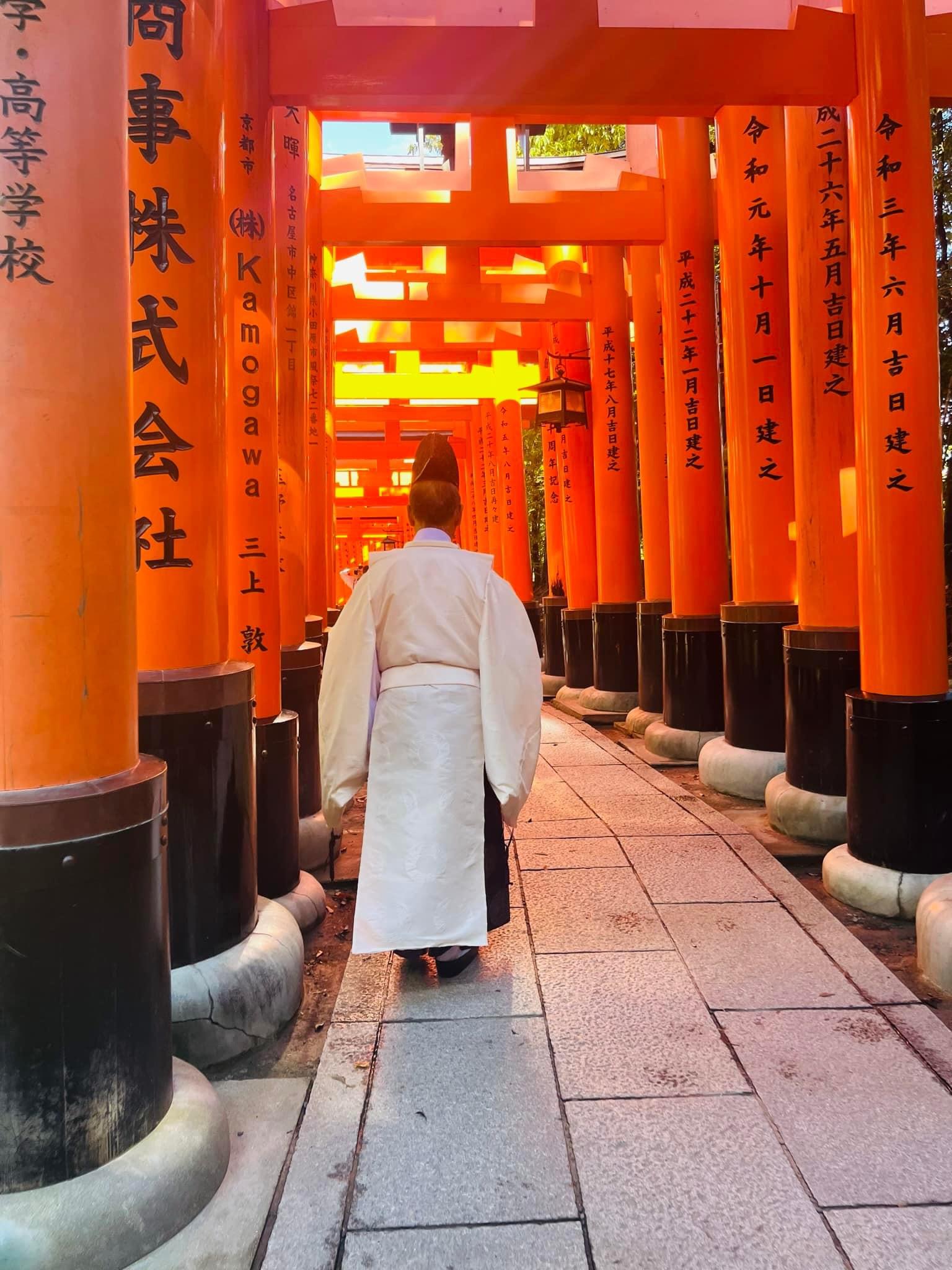 Buddhist priest at Fushimi Inari. Large red tori gates with black Japanese characters create a covered pathway that a Buddhist priest in white robe walks.