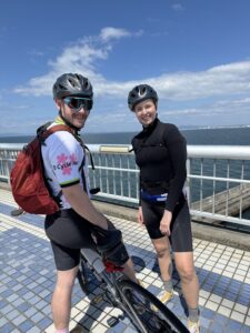 Two cyclist look at camera while standing on a bridge with the ocean in the back ground on a blue sky day.