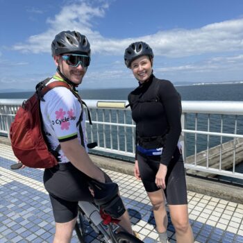 Two cyclist look at camera while standing on a bridge with the ocean in the back ground on a blue sky day.