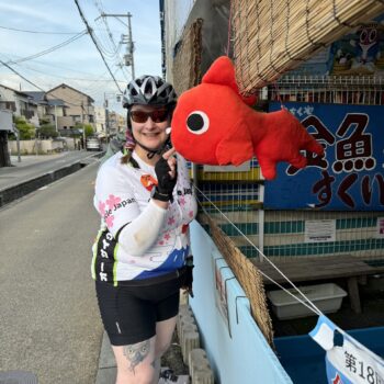 Cyclist gets close to red stuffed fish hanging from a storefront.