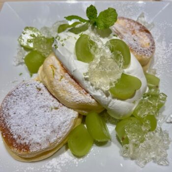 A delicious display of Japanese fluffy pancakes covered with white icing sugar a huge dollop of whip cram and a green fruit beside and on the top of the whip cream.