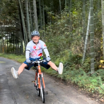 Man coasts on bicycle with feet up in the air as he rides on a paved pathway through a bamboo grove.