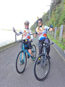 Two cyclists give a thumbs up as they pose with their bikes on a pathway next to the Ocean in Kansai, Japan.