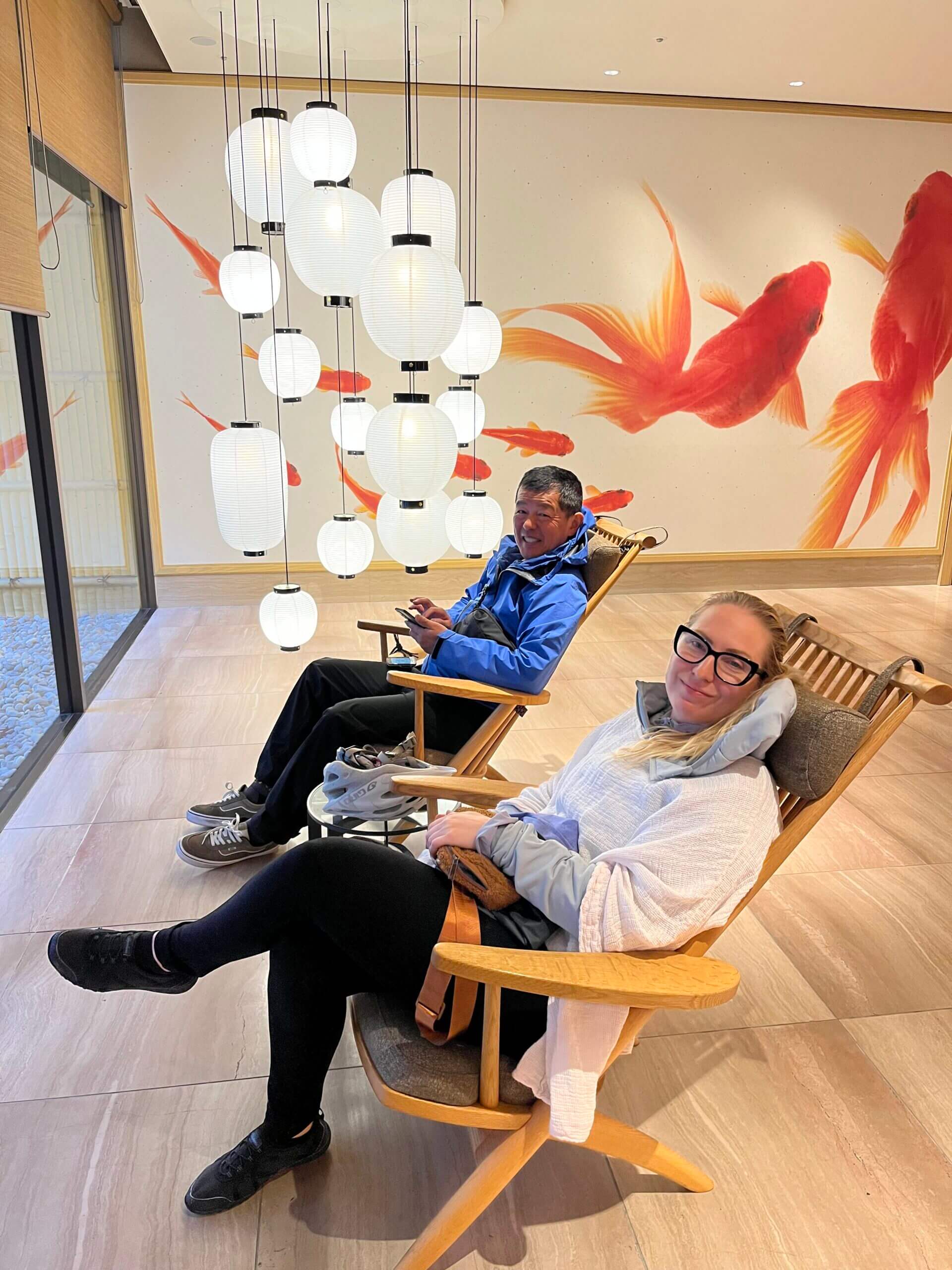 Two people sit in lounge chairs in hotel lobby. White lanterns hang from ceiling and large gold fish are painted on back wall.